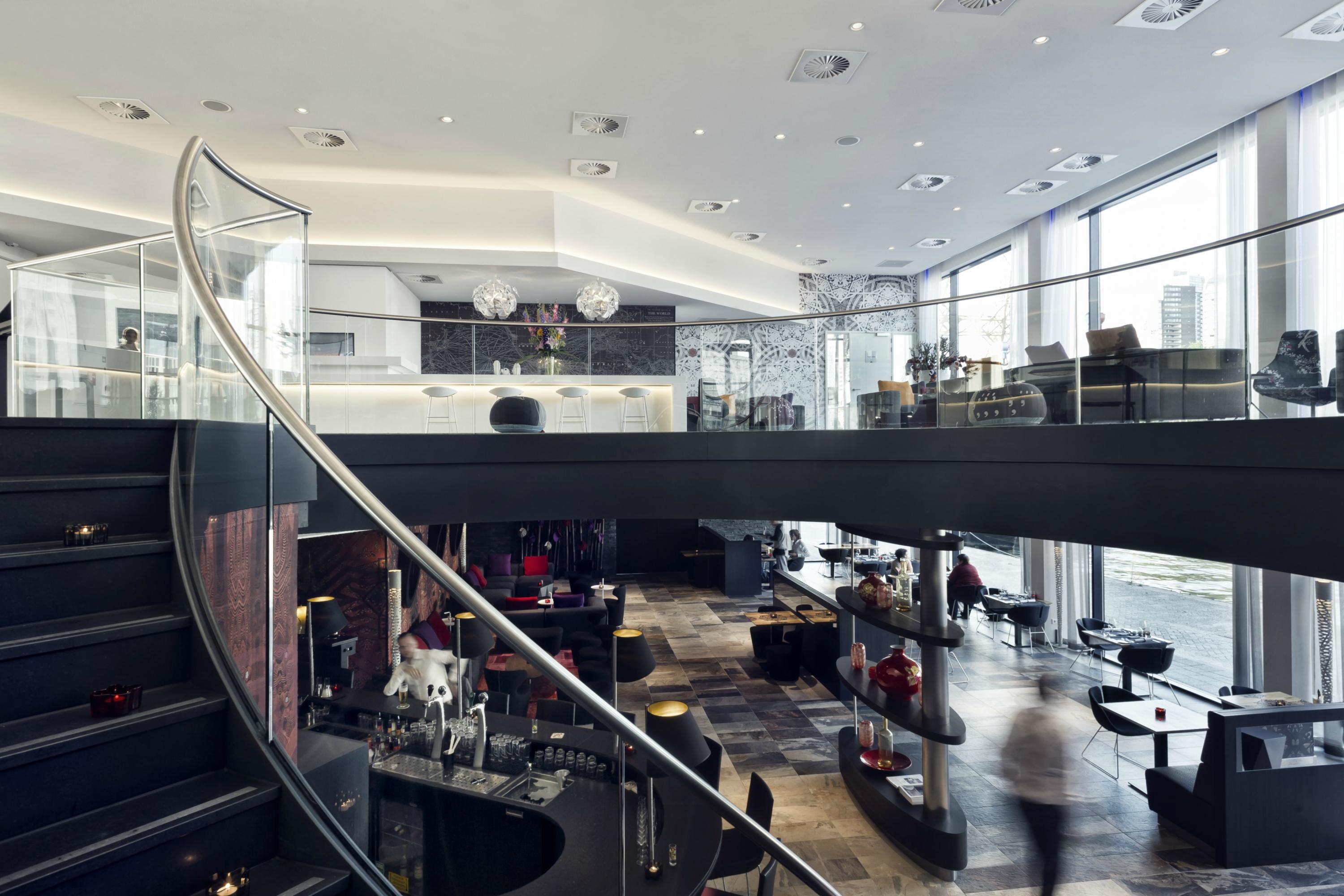 Mainport Hotel Rotterdam, A Hilton Affiliate Hotel Εξωτερικό φωτογραφία The photo shows a modern restaurant or lounge area with a sleek and sophisticated design. It features a large, open space with high ceilings and large windows that allow natural light to flood in. A curved staircase leads to a second level, creating 