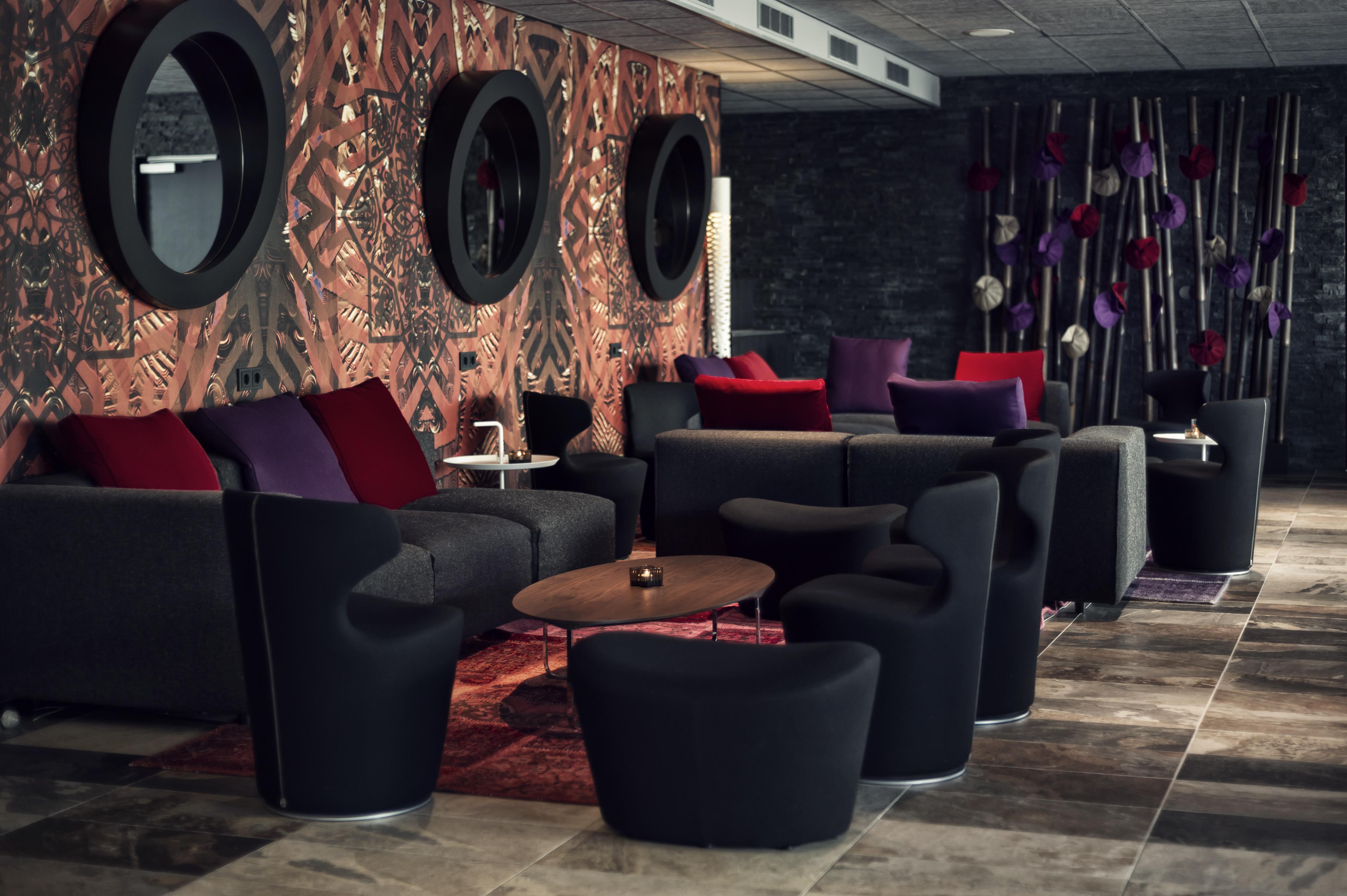 Mainport Hotel Rotterdam, A Hilton Affiliate Hotel Εξωτερικό φωτογραφία The photo shows a modern lounge area featuring various seating arrangements. There are several dark, contemporary chairs and sofas, some adorned with colorful cushions in shades of purple and red. The background has a patterned wall with round mirror