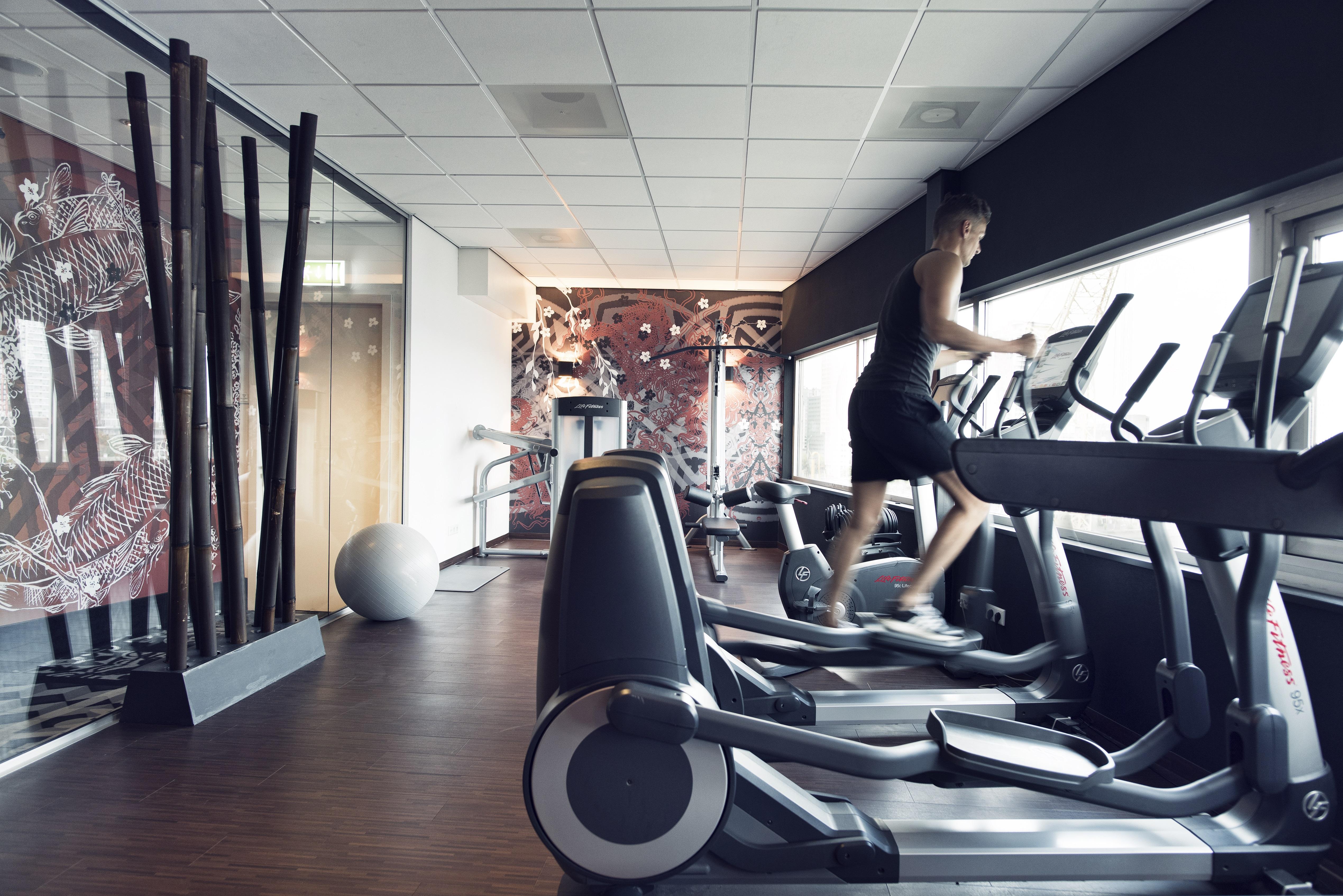 Mainport Hotel Rotterdam, A Hilton Affiliate Hotel Εξωτερικό φωτογραφία The photo shows a modern gym or fitness room. In the foreground, there are several exercise machines, specifically elliptical trainers, and a person is using one of them. The gym features a large window that likely provides natural light, and there i