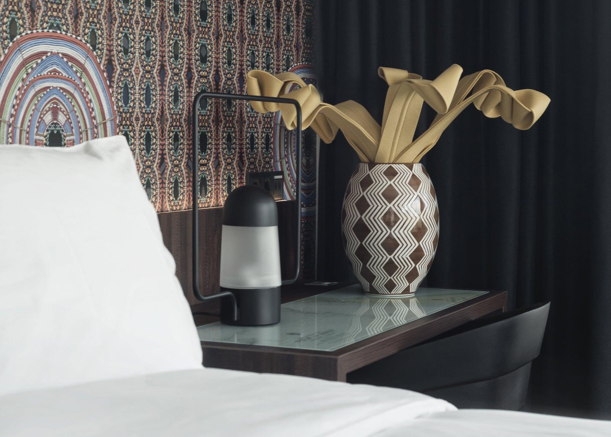 Mainport Hotel Rotterdam, A Hilton Affiliate Hotel Εξωτερικό φωτογραφία The photo shows a modern bedroom setting with a neatly made bed featuring white linens. Next to the bed is a stylish bedside table with a glass top and a patterned wooden base. On the table, there is a unique vase containing sculptural yellow decor r