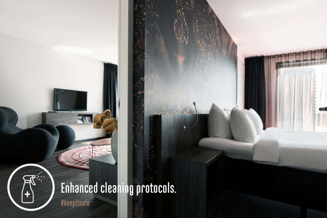 Mainport Hotel Rotterdam, A Hilton Affiliate Hotel Εξωτερικό φωτογραφία The photo shows a modern hotel room with a sleek and minimalist design. On one side, there is a bed with large, white pillows and a dark headboard featuring an abstract design. Next to the bed is a dark wooden nightstand. The other side of the room i